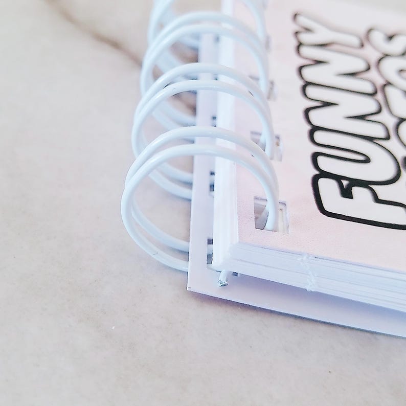 May include: A white spiral-bound notebook featuring a portion of the text "FUNNY" printed in bold black letters on the cover. The binding consists of white metal wire loops that secure the pages together along the left edge of the notebook.