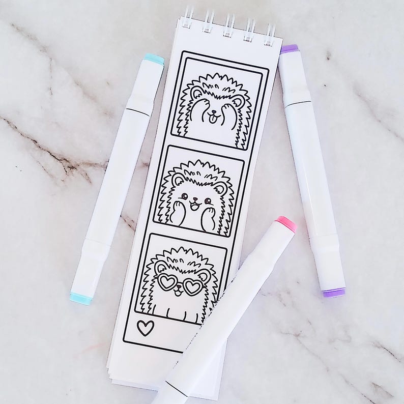 May include: A white spiral-bound notepad featuring three vertical black and white illustrations of a hedgehog in different poses, accompanied by three markers with light blue, pink, and purple caps positioned around the notepad.