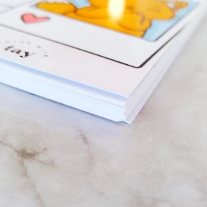 May include: A stack of white rectangular cards with a glossy finish. The top card features a cartoon illustration of an orange character, a small pink heart, and the text "COLOR WITH TAY" printed in black ink on the lower left corner of the card.
