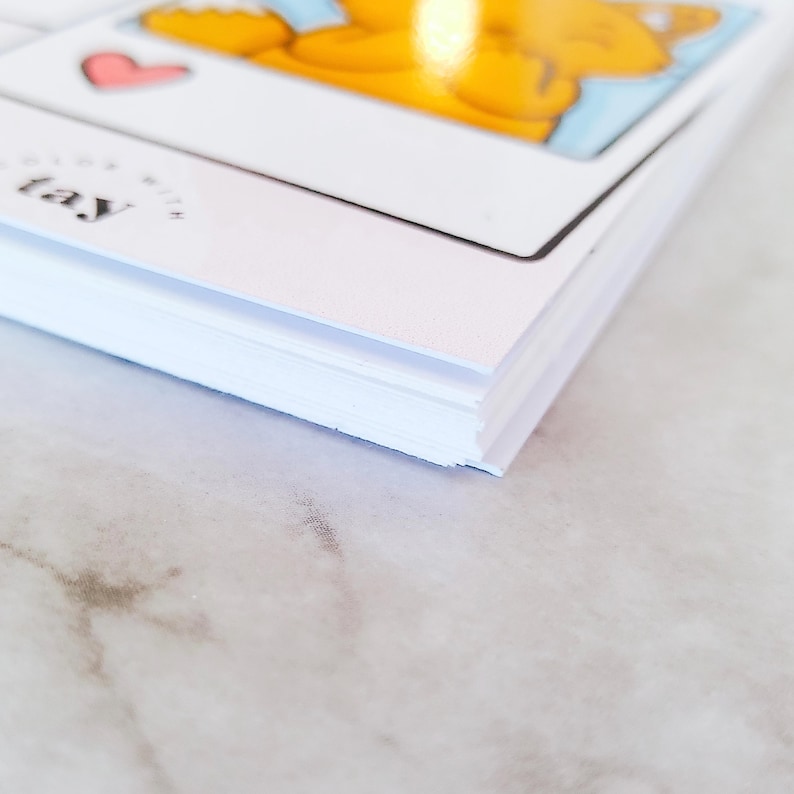May include: A stack of white rectangular cards with a glossy finish. The top card features a cartoon illustration of an orange character, a small pink heart, and the text "COLOR WITH TAY" printed in black ink on the lower left corner of the card.