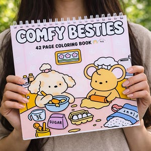 May include: A spiral-bound coloring book titled "COMFY BESTIES" with a pink cover. The cover features cartoon illustrations of a dog and a mouse cooking. The book contains 42 pages. The background is a soft pink with flower and heart patterns.
