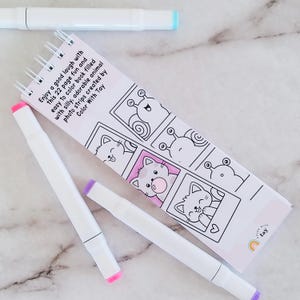 May include: A spiral-bound 22-page coloring book featuring black and white line art of animals in photo strips, with the text 'Enjoy a good laugh with this 22 page fun and easy to color book filled with silly, adorable animal photo strips created by Color With Tay'. Three white markers with colored caps are placed around the book.