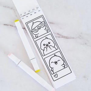 May include: A spiral-bound coloring pad featuring three vertical black and white illustrations of a small bird character in different poses, accompanied by two white markers with yellow and light pink caps respectively.