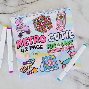 Op de afbeelding: Een spiraalgebonden kleurboek getiteld "Retro Cutie" met 42 pagina's, met illustraties van een rolschaats, een Rubik's kubus, een mobiele telefoon en andere retro-thema's. Inclusief twee stiften met gele en paarse punten.