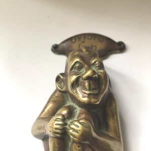 Peut inclure: Un heurtoir de porte vintage en laiton représentant une figure souriante et assise avec de grandes dents. Le heurtoir a un dessus incurvé avec le mot "Oxford" inscrit. Le laiton a un aspect vieilli et antique, avec des détails et des textures visibles.