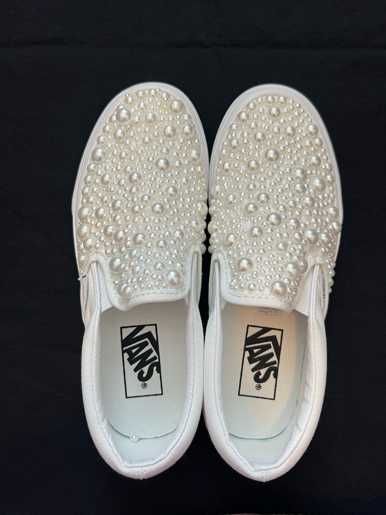 Puede incluir: Un par de zapatillas Vans blancas de deslizamiento cubiertas de adornos de perlas blancas. El logotipo de Vans es visible en el interior de cada zapato.