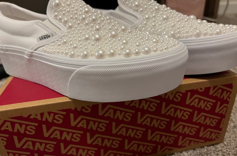 Puede incluir: Zapatillas Vans blancas de deslizamiento con suela de plataforma y cubiertas de perlas blancas. Los zapatos est&aacute;n sobre una caja con el logotipo de Vans repetido en rojo.