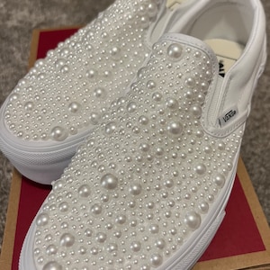 Puede incluir: Zapatillas Vans blancas de deslizamiento cubiertas de adornos de perlas blancas. Los zapatos est&aacute;n en una caja marr&oacute;n con el logotipo de Vans.