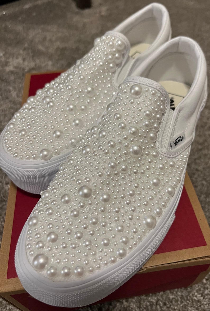 Puede incluir: Zapatillas Vans blancas de deslizamiento cubiertas de adornos de perlas blancas. Los zapatos est&aacute;n en una caja marr&oacute;n con el logotipo de Vans.
