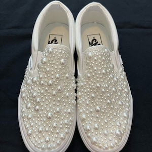 Puede incluir: Un par de zapatos de lona blancos con un adorno de perlas. Los zapatos tienen una suela de goma blanca y un logotipo de Vans negro en la parte posterior.