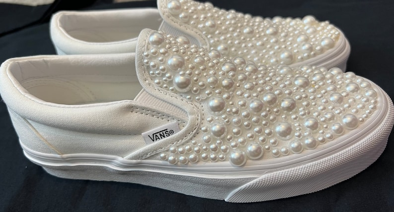 Puede incluir: Zapatillas Vans blancas de lona con cierre de slip-on y suela de goma blanca. Las zapatillas est&aacute;n cubiertas de cuentas blancas que parecen perlas. El logotipo de Vans es visible en el lateral de la zapatilla.