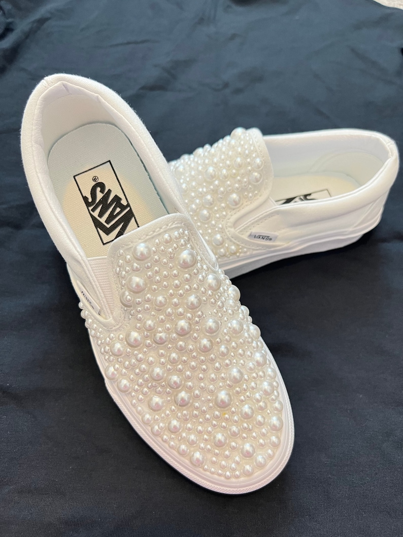 Puede incluir: Zapatillas Vans blancas de deslizamiento con un adorno de perlas en la parte superior. El logotipo de Vans es visible en el lateral del zapato.