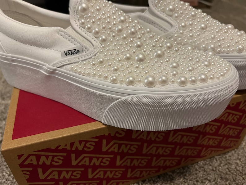 Puede incluir: Zapatillas Vans blancas de plataforma con deslizamiento y adorno de perlas. Las zapatillas est&aacute;n sobre una caja marr&oacute;n con el logotipo de Vans impreso.