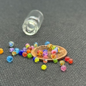 以下が含まれることがあります： 青、オレンジ、ピンク、緑など、さまざまな色のミニチュアガラス玉が暗い表面に散らばっています。小さな透明なガラスバイアルも存在します。ビー玉はペニーの隣にあります。