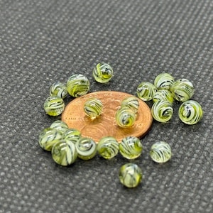 May include: A collection of small, round glass beads with a swirl pattern of white and yellow-green. The beads are scattered around a copper-colored penny on a textured, dark gray surface. The beads are approximately 0.5 inches in diameter.
