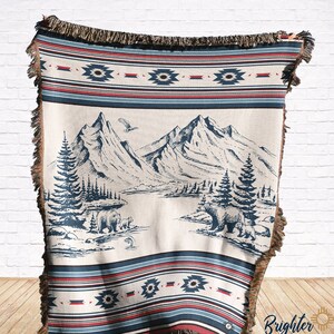 May include: A woven throw blanket with a cream background featuring a blue and white mountain scene with bears and trees. The blanket has a red, blue, and white geometric border.