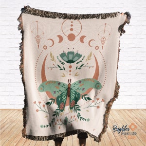 May include: A woven tapestry throw blanket with a cream background featuring a green butterfly with floral accents and a moon and sun design. The throw blanket has a fringe border.
