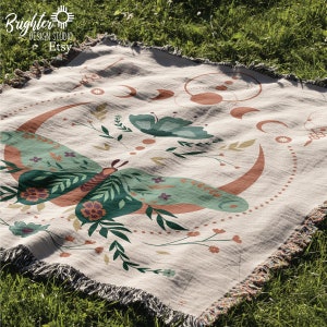 May include: A woven throw blanket with a cream background and a colorful design featuring a butterfly, crescent moons, and floral elements. The text "Brighter Design Studio Etsy" is printed in the top left corner.