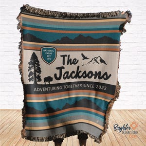 May include: A woven throw blanket with a tan background and blue, brown, and orange stripes. The blanket features a mountain scene with a stylised eagle and the text "The Jacksons Adventuring Together Since 2022". A small circle with the text "National Parks Est 1872" is in the top left corner.