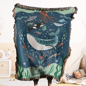 May include: A blue and green woven throw blanket with a whale, octopus, and other sea creatures. The whale has the name "Austin" written on it. The blanket also has the date "1.27.23" and the numbers "34.445825" and "118.077936".