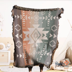 May include: A woven throw blanket with a geometric pattern in shades of brown, orange, and green. The blanket has a fringe border and is made of a soft, cosy material.