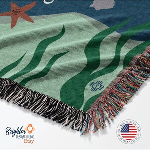 May include: A woven throw blanket with a green and blue abstract design featuring a starfish, waves, and a small flower. The blanket has a fringe border and is made in the USA.