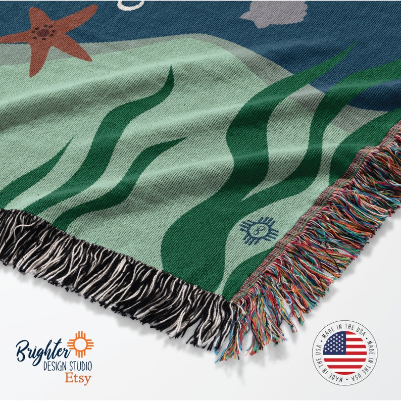 May include: A woven throw blanket with a green and blue abstract design featuring a starfish, waves, and a small flower. The blanket has a fringe border and is made in the USA.