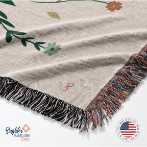 May include: A woven throw blanket with a floral pattern in shades of green, brown, and orange. The blanket has a black fringe border and a label that says "B". The blanket is made in the USA.