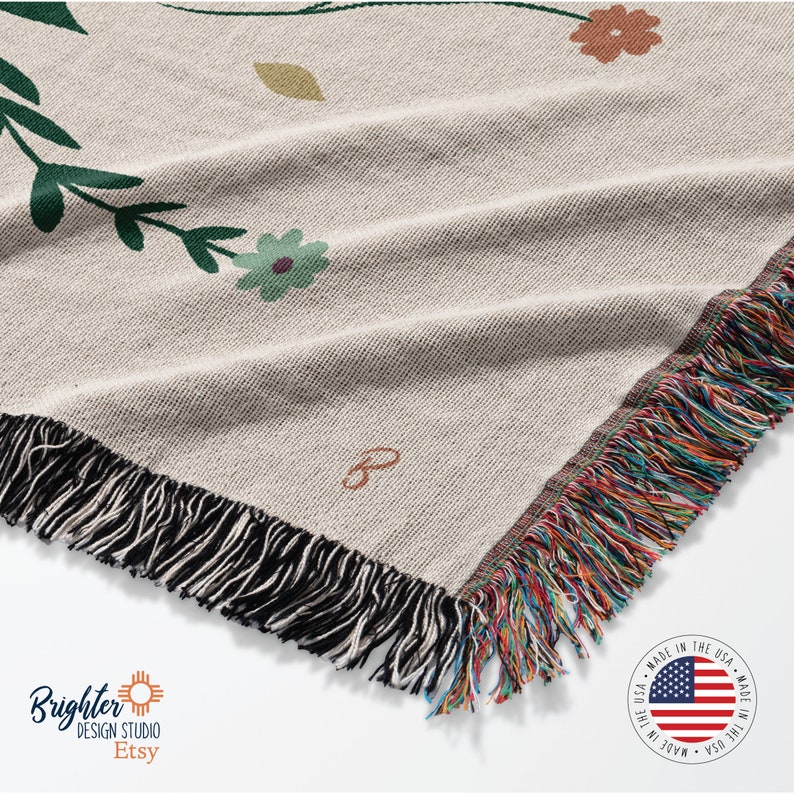 May include: A woven throw blanket with a floral pattern in shades of green, brown, and orange. The blanket has a black fringe border and a label that says "B". The blanket is made in the USA.