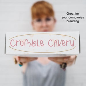 K&ouml;nnte beinhalten: Eine wei&szlig;e B&auml;ckereibox mit dem Schriftzug "Crumble Cakery" in rosa Schrift. Die Box hat einen goldenen ovalen Rahmen mit kleinen Herzakzenten. Die Box wird von einer Person im Hintergrund gehalten.