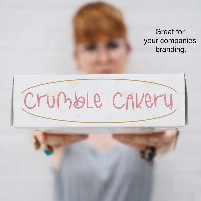 K&ouml;nnte beinhalten: Eine wei&szlig;e B&auml;ckereibox mit dem Schriftzug "Crumble Cakery" in rosa Schrift. Die Box hat einen goldenen ovalen Rahmen mit kleinen Herzakzenten. Die Box wird von einer Person im Hintergrund gehalten.