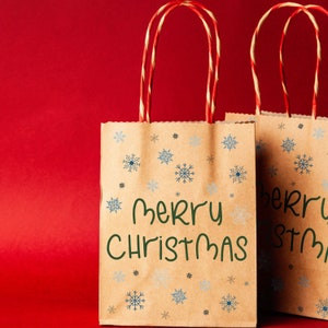 K&ouml;nnte beinhalten: Zwei braune Geschenkpapier-Taschen mit rot-wei&szlig; gestreiften Griffen. Die Taschen sind mit blauen Schneeflocken und den Worten "Merry Christmas" in Gr&uuml;n verziert. Der Hintergrund ist einfarbig rot.