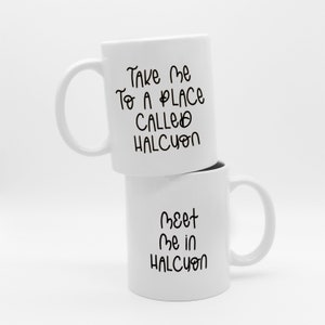 K&ouml;nnte beinhalten: Zwei wei&szlig;e Keramikbecher, die &uuml;bereinander gestapelt sind. Der obere Becher hat den Text "Take Me To A Place Called Halcyon" in schwarzer Schrift. Der untere Becher tr&auml;gt die Aufschrift "Meet Me in Halcyon". Beide Becher haben einen gebogenen Griff.