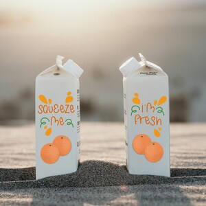 K&ouml;nnte beinhalten: Zwei wei&szlig;e Saftkartons mit orangefarbenen und gr&uuml;nen Grafiken und den S&auml;tzen "squeeze me" und "I'm fresh". Jeder Karton zeigt ein Paar Orangenillustrationen. Die Kartons stehen auf einer sandigen Oberfl&auml;che.