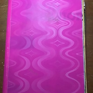 May include: A hot pink binder with a repeating wavy pattern in a lighter shade of pink. The binder is closed and has a spine on the left side. The background is a wooden surface.