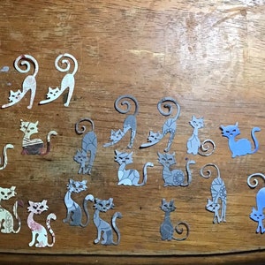 May include: A collection of cat-shaped paper cutouts in various colors and patterns, including white, gray, and blue. The cats are in different poses, some with curled tails, arranged on a wooden surface. The cutouts vary in size and design.