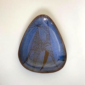 May include: A triangular ceramic dish with a blue glazed finish and a brown rim. The interior features a dark brown line drawing of a tall, tower-like structure with a radiating pattern at the top and a solid brown shape at the base.