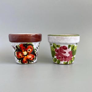 May include: Two small, hand-painted ceramic flower pots. One pot has a brown rim and a large orange and red flower design. The other pot has a white base with a green rim and a red rose design.