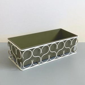 May include: Olive green rectangular planter with a white metal decorative frame. The frame has a repeating, curved design. The planter sits on a light grey surface against a white background. Ideal for plants.