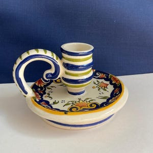 May include: A white ceramic candle holder with a blue and green striped candle cup and a handle. The base of the holder is decorated with floral designs in blue, yellow, red, and green.