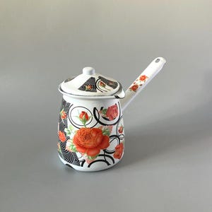 May include: A white enamel coffee pot with a long handle and a lid, featuring a decorative pattern of vibrant orange roses and black circular geometric lines. The design covers the body of the pot and the lid, with smaller rose accents on the handle.