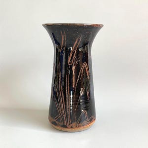 May include: A dark brown ceramic vase with a flared top. The vase has a glossy finish and is decorated with etched, stylized plant-like designs in a lighter brown colour. The base of the vase has a lighter, unglazed appearance.