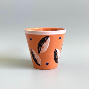 May include: A terracotta-coloured ceramic cup featuring a white rim and a hand-painted pattern of dark brown and light beige almond-shaped motifs with wavy lines, accented by small, scattered dark blue dots across the exterior surface.