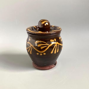 May include: A small, dark brown ceramic jar with a lid. The jar is decorated with a yellow floral design. The lid has a small knob on top. The jar sits on a reddish-brown base. A decorative kitchen item.