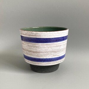 May include: A ceramic bowl featuring a wide, flared rim and a tapered base. The exterior displays horizontal stripes in deep blue and textured white with brown undertones. The interior of the bowl is finished in a solid, smooth green glaze.