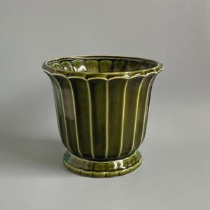 May include: A dark olive green ceramic planter with a glossy finish. The vessel features a vertical ribbed or fluted pattern along its exterior body, a wide flared rim with a scalloped edge, and a circular pedestal base for stability.