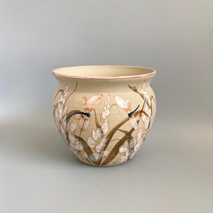 May include: A round ceramic planter with a beige base colour, featuring hand-painted illustrations of dragonflies and tall grasses in shades of brown, white, and soft orange. The pot has a wide, flared rim with a thin reddish-brown painted edge.