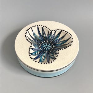 May include: A round ceramic trinket dish with a floral design. The dish features a cream-coloured base with a blue and black floral pattern in the centre. The rim is light blue with black detailing. The dish is suitable for holding small items.