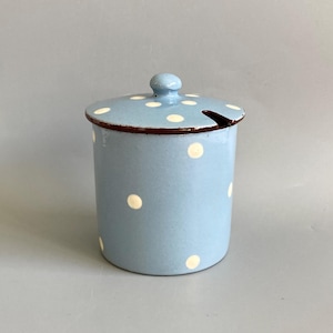 May include: A light blue cylindrical canister with a matching lid featuring white polka dots. The container has a dark brown rim and a small notch on the lid edge for a spoon. The surface has a smooth, glossy finish with a small knob on the lid.
