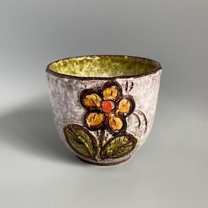 May include: A ceramic cup with a textured, mottled light grey exterior and a vibrant lime green interior. The front features a raised, embossed flower design with yellow petals, a bright orange centre, and dark green leaves outlined in dark brown.